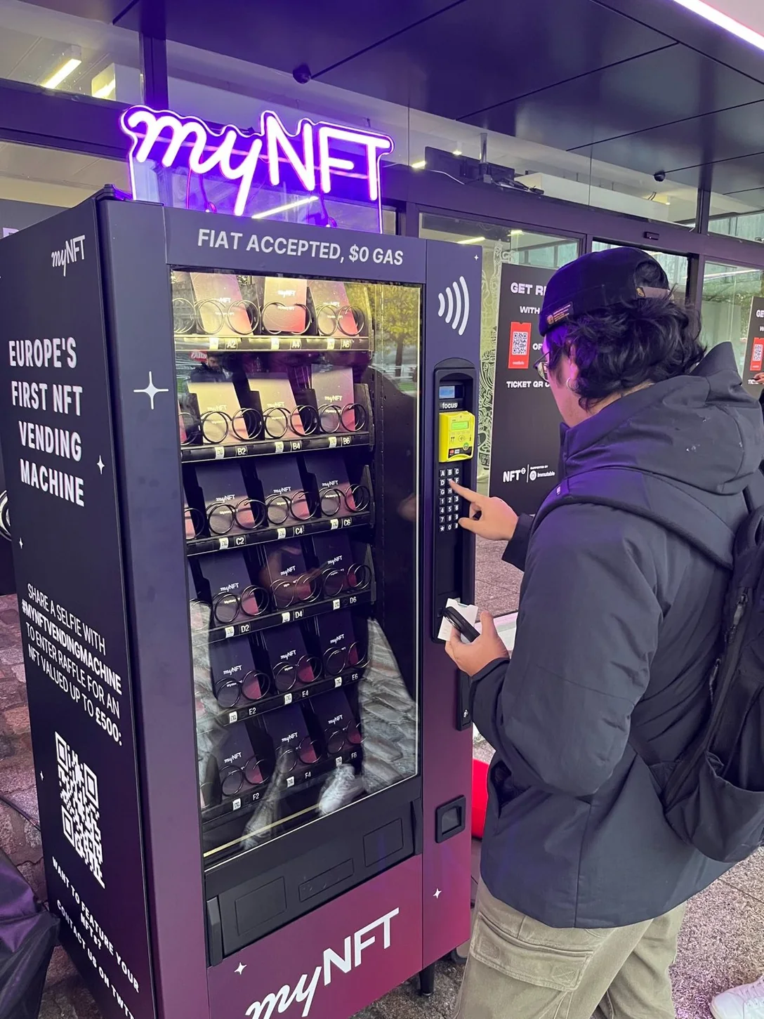 A visitor pressing the keypad of the myNFT vending machine outside the NFT.London conference at dusk. The machine is magenta and black under a lit neon myNFT sign, printed with “Europe’s first NFT vending machine” and “Fiat accepted, $0 gas”, its racks stacked with myNFT-branded packages.