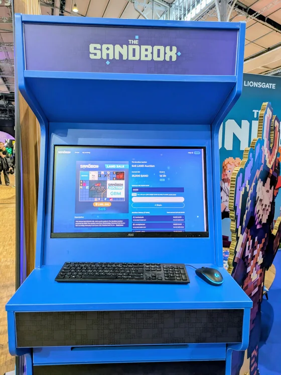 A blue arcade cabinet branded The Sandbox, its screen showing a 6x6 LAND auction running on GBM with a current bid of 35,000 SAND and a line reading “You will earn 2,931.42 SAND (5.43%) if outbid”.